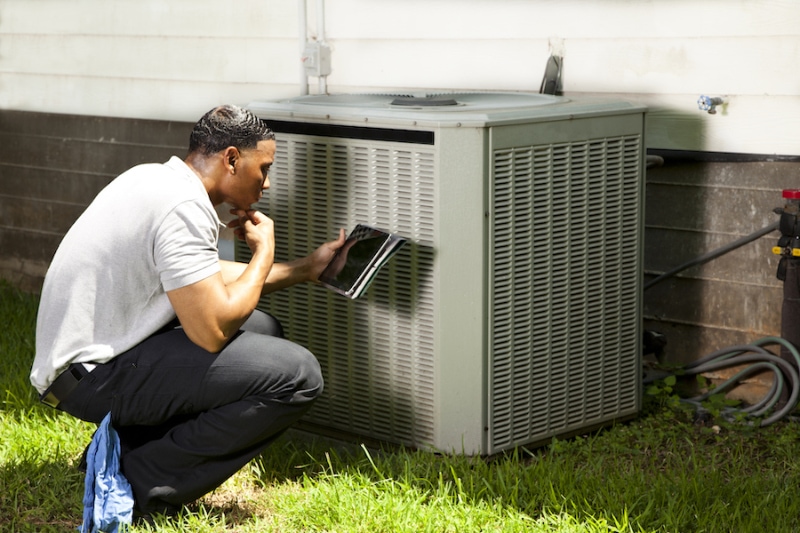 Home inspector, repairman, adjuster examines air conditioner units at a customer home. He is using his digital tablet to record results.