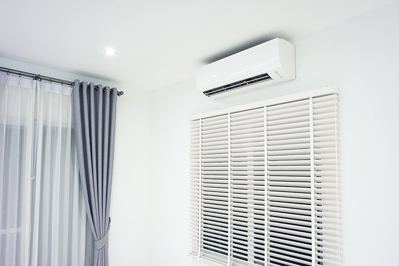 A modern mini-split air conditioner unit mounted on a wall above window blinds in a bright room.